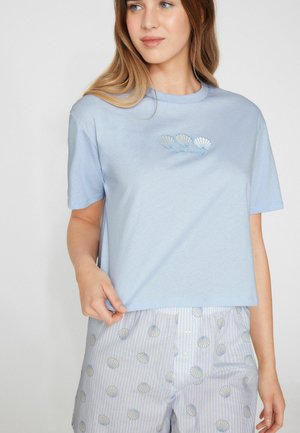 Frau trägt ein hellblaues Kurzarmshirt mit drei Muscheln und dem Schriftzug „das Meer ruft“, kombiniert mit hellen Hosen mit Muschelmuster.