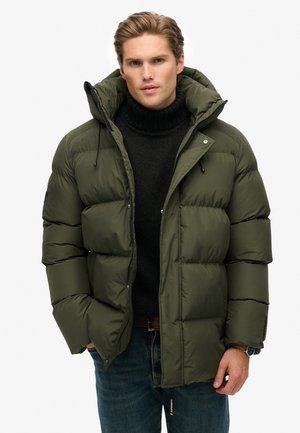 Olive green puffer jacket with a hood, featuring a zip closure, large quilted sections, and black accents, worn over a dark turtleneck.