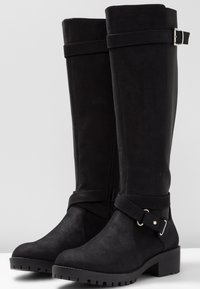 Botas negras hasta la rodilla hechas de ante, con una cremallera lateral, una correa decorativa con una hebilla plateada y una suela de goma texturizada.