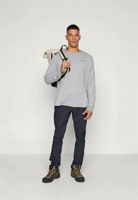 Jasnoszara koszula z długim rękawem z małym logo, w połączeniu z ciemnonawigacyjnymi spodniami i czarnymi butami trekkingowymi z złotymi akcentami. Niesie beżowy plecak.