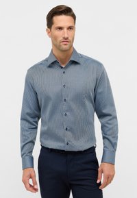 Uomo che indossa una camicia a maniche lunghe blu aderente con un motivo, abbinata a pantaloni blu scuro, in piedi davanti a uno sfondo neutro.