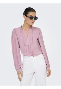 Blouse longue rose pâle à manches longues avec un décolleté en V, des volants, des détails plissés, associée à un pantalon blanc et des lunettes de soleil rondes sombres.