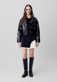 Veste en cuir synthétique noire avec des boutons dorés, haut noir ajusté, jupe plissée asymétrique noire et crème, et bottes noires montantes.