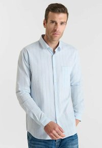Man met kort bruin haar in een lichtblauw overhemd met dunne verticale strepen en blauwe spijkerbroek, staand tegen een effen achtergrond.