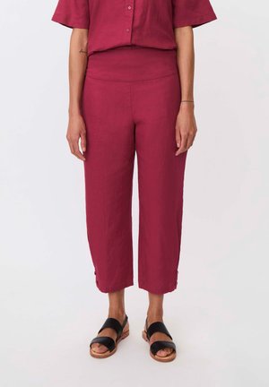 Kvinde iført korte, højttaljede, løstsiddende mørkerøde bukser med knapdetaljer ved kanten og sorte åbne sandaler.