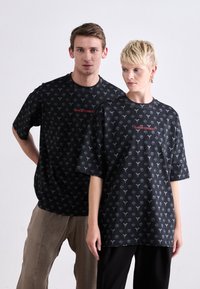 Sorte grafik t-shirts med et gentaget mønster af symboler i grå. Centreret rød tekst læser "CARLO COLECCI." Løs pasform og korte ærmer.