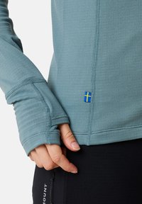 Light blue long-sleeve top with thumb holes, textured fabric, contrasting stitching, and a small Swedish flag patch near the hem.