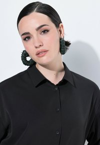 Schwarzes Hemd mit Knöpfen und glatter Textur, ausgestattet mit einem Kragen und sichtbaren Knöpfen. Das Modell trägt große, grüne Haargummie-Ohrringe im Scrunchie-Stil.