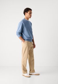 Ανοιχτό μπλε φούτερ με πλεχτές μανσέτες και στρίφωμα, σε συνδυασμό με μπεζ παντελόνι. Το μοντέλο στέκεται προφίλ, φοράει άσπρα παπούτσια με καφέ σόλες.