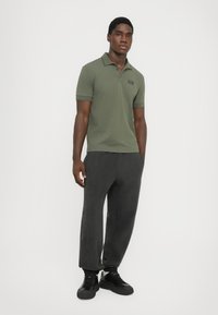 Olive green polo shirt with a slim fit and black logo, paired with dark grey textured joggers and black lace-up sneakers.