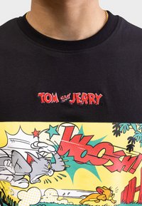 Melnas kokvilnas T-krekls ar izšūtu uzrakstu "TOM un JERRY" sarkanā krāsā. Grafiskais prints dzeltenā krāsā ar multfilmu varoņiem Tomu un Džeriju.