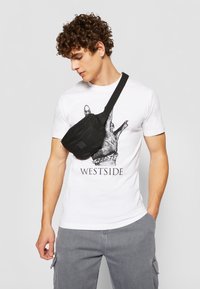 Sac en toile noire avec fermeture éclair, porté en diagonale sur le corps, sur un t-shirt blanc arborant un graphisme de main et le texte « WESTSIDE ». 