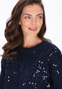 Vrouw met golvend bruin haar, gekleed in een marineblauwe trui versierd met reflecterende sequins, die iets opzij kijkt en zachtjes glimlacht.
