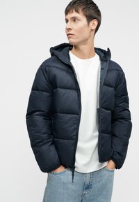 Chaqueta puffer azul marino con capucha, cierre de cremallera y diseño acolchado. Llevada sobre una camiseta blanca y jeans de mezclilla azul claro.