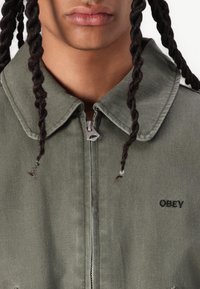 Grønn denimjakke med glidelås, stor krage og brodert "OBEY"-logo. Har et strukturert stoff og flettet hår.