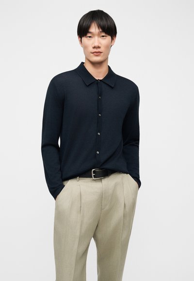 Νέος άνδρας που φοράει μαύρο μακρυμάνικο πουκάμισο με κουμπιά, μπαίνει μέσα σε μπεζ πλισέ παντελόνια, με τα χέρια στις τσέπες, στέκεται μπροστά σε λευκό φόντο.