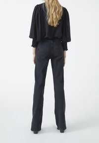 Schwarze Wide-Leg-Jeans mit hoher Taille, aus Denim gefertigt. Verfügt über zwei Hintertaschen und einen sauberen Saumabschluss. Das Modell trägt eine dunkle Bluse.