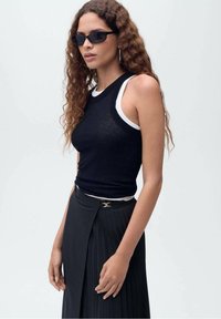 Un haut sans manches noir superposé sur un débardeur blanc, accompagné d'une jupe plissée noire avec une clasp décorative. Le mannequin porte des lunettes de soleil noires.