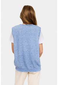 Pulover fără mâneci, tricotat, de culoare albastră deschisă, cu un tiv cu nervuri, având un finisaj texturat. Purtați peste un tricou alb, expunând umărul și spatele.