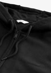 Veste à capuche zippée noire avec un intérieur doux, dotée d'une capuche à cordon de serrage, d'une fermeture éclair et d'un tissu texturé. L'étiquette à l'intérieur indique "NEXT."