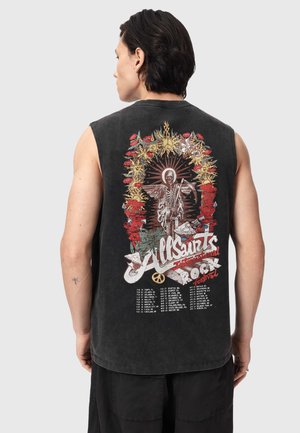 Person mit kurzem dunklem Haar, trägt ärmelloses schwarzes Shirt mit farbenfrohem Skelettmotiv, Blumengirlande und Text mit Auflistung von Musikfestival-Daten.