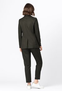 Tmavě zelený oblek s přizpůsobeným sakem a slim-fit kalhotami, s jedním zadním rozventilováním. Nosený s bílými teniskami pro kontrast.