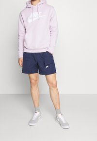 Svetle fialová mikina s kapucí a bílým logem Nike, spárovaná s námořnickými šortkami s bočními kapsami a šedými botami Nike s bílou podešví a světle šedými ponožkami.