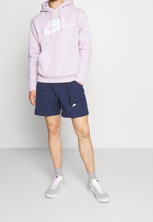 Svetle fialová mikina s kapucí a bílým logem Nike, spárovaná s námořnickými šortkami s bočními kapsami a šedými botami Nike s bílou podešví a světle šedými ponožkami.