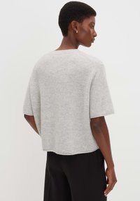 Femme portant un pull en laine gris clair à manches courtes et un pantalon noir, vue de dos et tournée sur le côté.