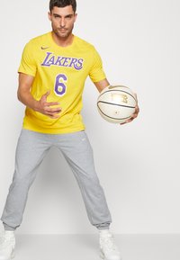 Tricou galben cu grafică purpurie "Lakers" și numărul 6, combinat cu pantaloni de trening gri. Ținând o minge de baschet, purtând adidași albi.
