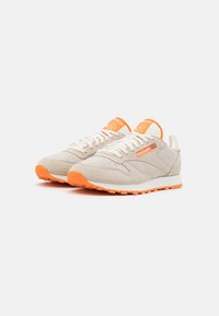 Chaussures de sport en daim gris clair avec des accents orange. Présentent une surface texturée, des lacets blancs et une semelle en caoutchouc orange.