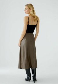 Schwarzes, enganliegendes Tanktop kombiniert mit einem hochtaillierten, braunen, kunstledernen Midirock. Das Modell trägt knielange, schwarze, spitze Stiefel.