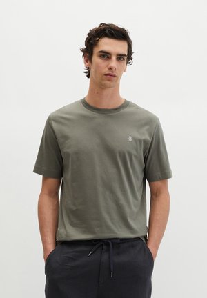 Junger Mann mit dunklen lockigen Haaren, trägt ein olivgrünes T-Shirt mit Rundhalsausschnitt und schwarze Hose mit Kordelzug, steht vor einem einfachen weißen Hintergrund.