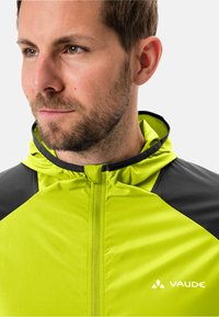 Limonengrüne Jacke mit einem schwarzen Schulterpanel, Reißverschluss und Kapuze. Leichter Stoff mit einem subtilen strukturierten Muster. Vaude-Logo sichtbar.
