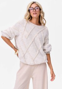 Kobieta z blond włosami, nosząca duże okulary, teksturowany jasnobeżowy sweter z podwiniętymi rękawami i jasnobeżowe spodnie, pozująca z jedną ręką na biodrze.