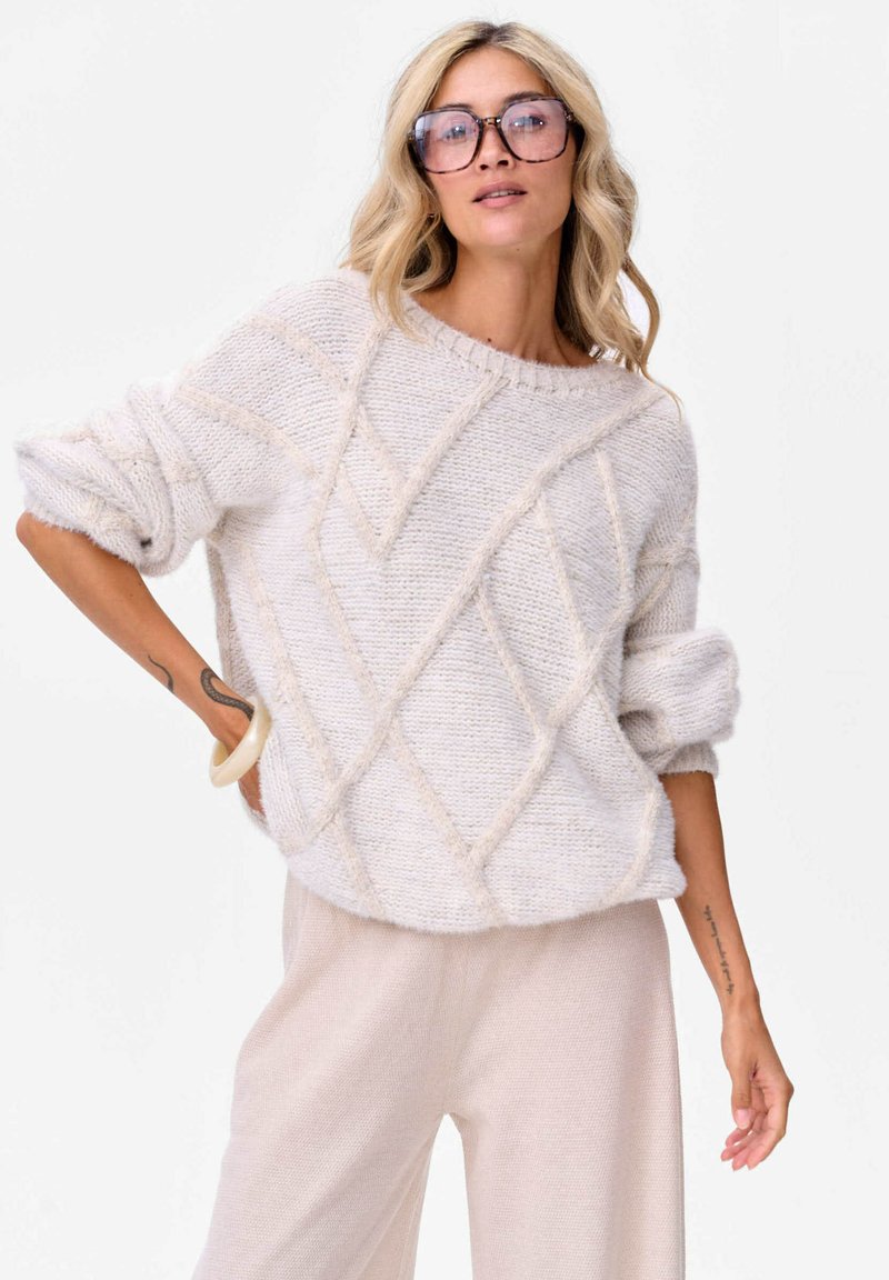 Kobieta z blond włosami, nosząca duże okulary, teksturowany jasnobeżowy sweter z podwiniętymi rękawami i jasnobeżowe spodnie, pozująca z jedną ręką na biodrze.