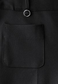 Un tissu noir au toucher lisse présente une poche rectangulaire avec un détail en anneau circulaire argenté au-dessus. Une couture simple souligne le design.