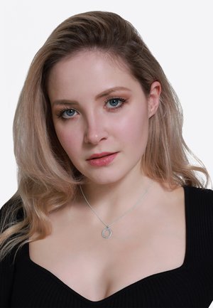 Collier en argent avec un pendentif circulaire orné de petites pierres précieuses, porté sur un haut noir à côtes, avec des vagues douces dans des cheveux de couleur claire.