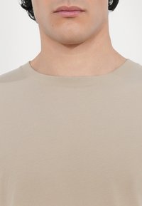 Gros plan sur la partie inférieure du visage et le cou d'une personne portant un t-shirt beige à col rond sur un fond uni.