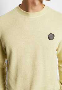 Jasnożółty sweter o fakturowanym splocie, z okrągłym dekoltem i ciemną, okrągłą naszywką z logo po lewej stronie klatki piersiowej.