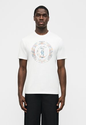 Άνδρας με λευκό μπλουζάκι που έχει πολύχρωμο κυκλικό λογότυπο και μαύρο παντελόνι, στέκεται μπροστά σε απλό λευκό φόντο.