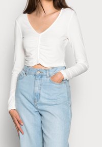 Valkoinen pitkähihainen crop-top, jossa on V-pääntie ja poimuttumia edessä, yhdistettynä korkeavyötäröisiin vaaleansinisiin farkkuihin.