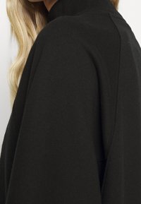 Cheveux blonds sur l’épaule portant un vêtement noir à manches longues avec un col rond haut, gros plan sur l’épaule et le haut du dos.