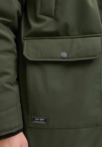 Olijfgroene jas met een gestructureerd oppervlak, voorzien van een voorkantvak en een ronde logoknop. Inclusief een label met merkinformaties.