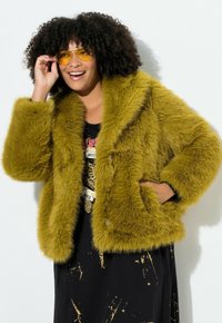 Faux fur jakke i olivengrøn, med en bred krave, to forlommer og en løs pasform. Båret over en sort kjole med guldsplatter.