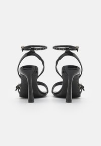Black high-heeled sandals feature shiny leather, ankle straps with gold studs, and a delicate bow accent at the back.