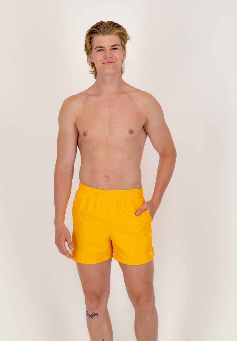 Geltoni maudymosi šortai su elastingu liemeniu, su šonu kišene ir lygiu audinio paviršiumi. Modelis stovi prieš paprastą foną.