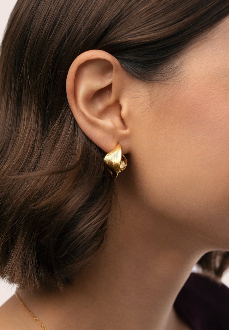Orecchini placcati in oro con design a torsione scanalata; finitura liscia e forma arrotondata; indossati su un modello con capelli corti.
