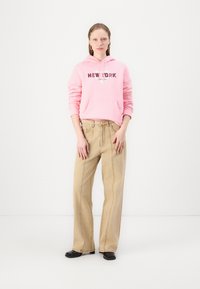 Felpa rosa con il testo "NEW YORK" e tasca a marsupio, abbinata a jeans beige a gamba larga e scarpe nere. Design semplice, materiale in cotone.