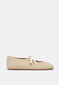 Beige suede ballet flat with rounded toe, cream-colored knotted ribbon lace on top, and textured rubber sole.